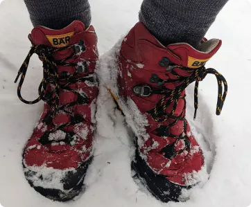 Rote Wanderschuhe mit Schnee bedeckt, getragen von einer Person mit grauen Socken auf verschneitem Boden.