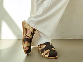 Damenfüße in stilvollen, braun-beigen Ledersandalen, weiße Hose, modernes Design