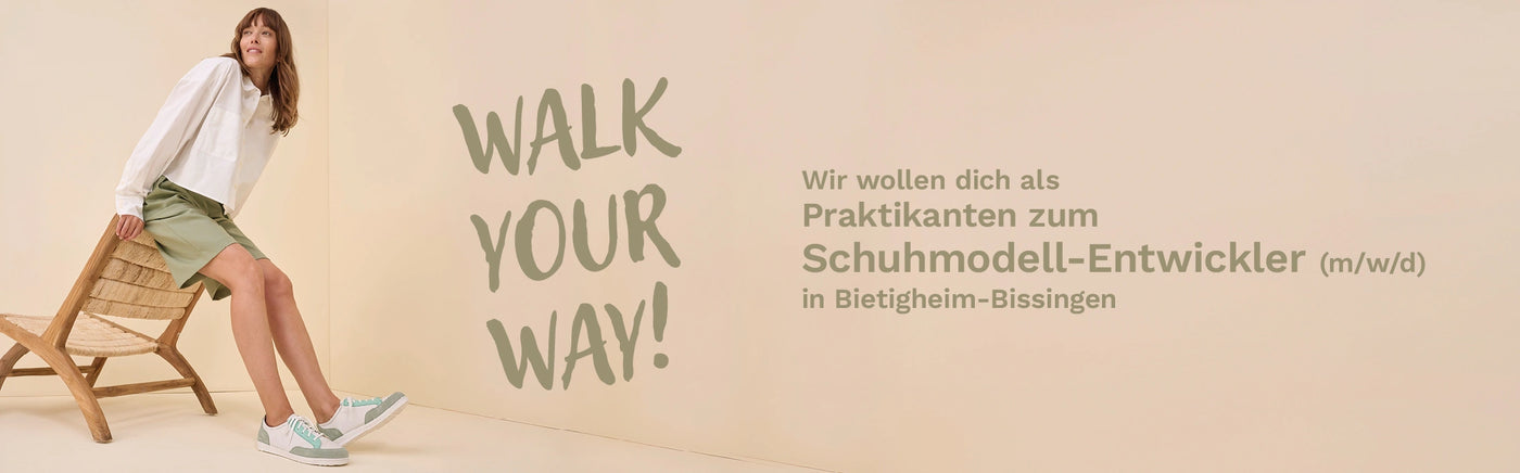 Vier Personen in Arbeitskleidung in einer Schuhmanufaktur stehen lächelnd vor Regalen mit Schuhen.