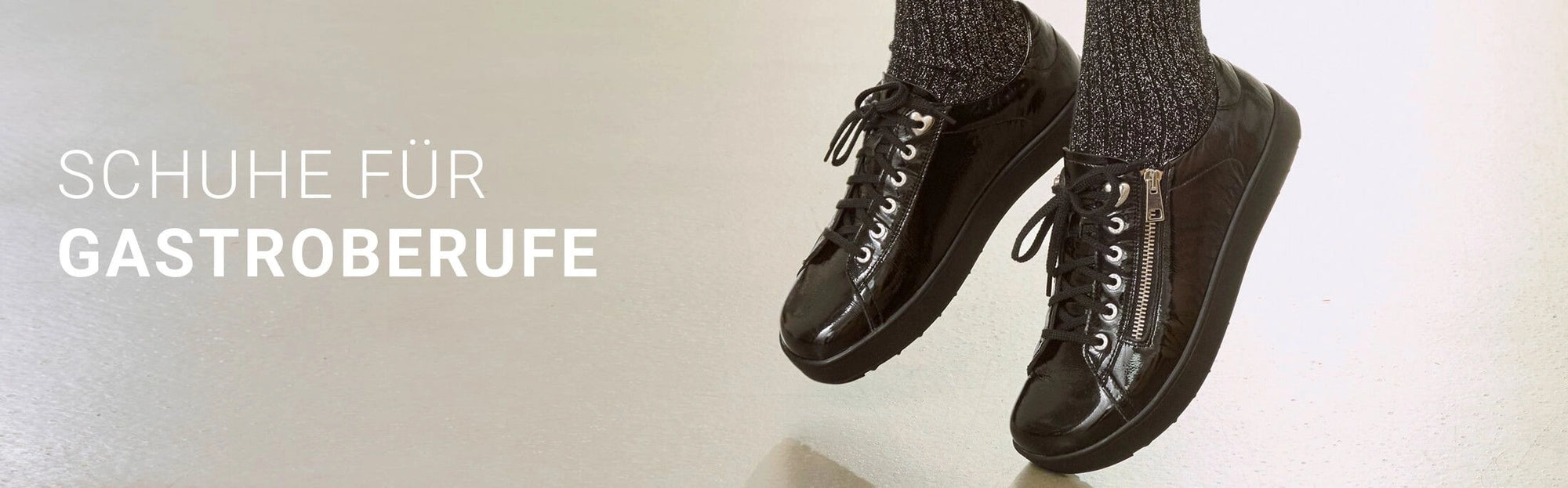 Werbebanner für Schuhe für Gastronomieberufe: Zwei schwarze, glänzende Damenschuhe mit Schnürsenkeln und seitlichem Reißverschluss sind an den Füßen einer Frau mit schwarzen Socken zu sehen. Links steht der Text ‚Schuhe für Gastroberufe‘