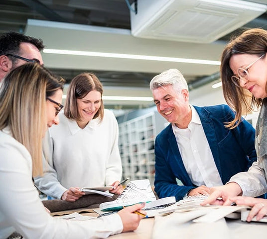 Eine Gruppe von Mitarbeitenden in einem Büro, die über einem Tisch voller Designunterlagen diskutieren und gemeinsam an einem Projekt arbeiten.