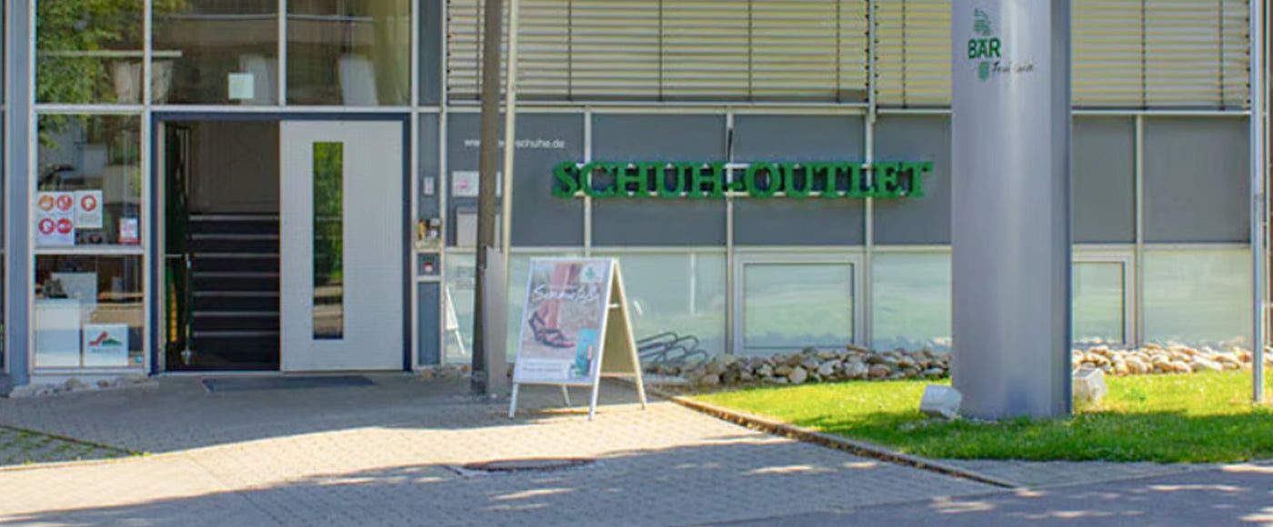 Eingangsbereich eines Schuhoutlets mit großen Glasfenstern und einem grünen Schild über der Tür.