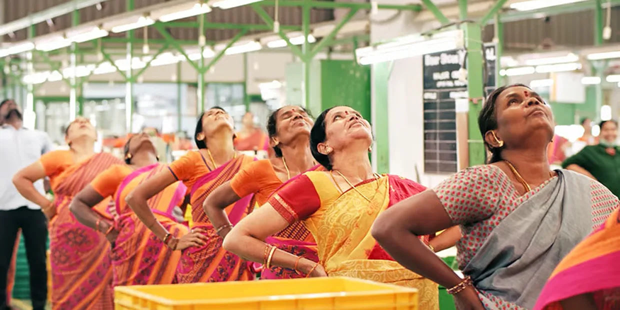 Mehrere Frauen in farbenfrohen Saris machen Dehnübungen in einer Fabrikhalle. Sie stehen in einer Reihe und strecken den Oberkörper und den Kopf nach hinten.