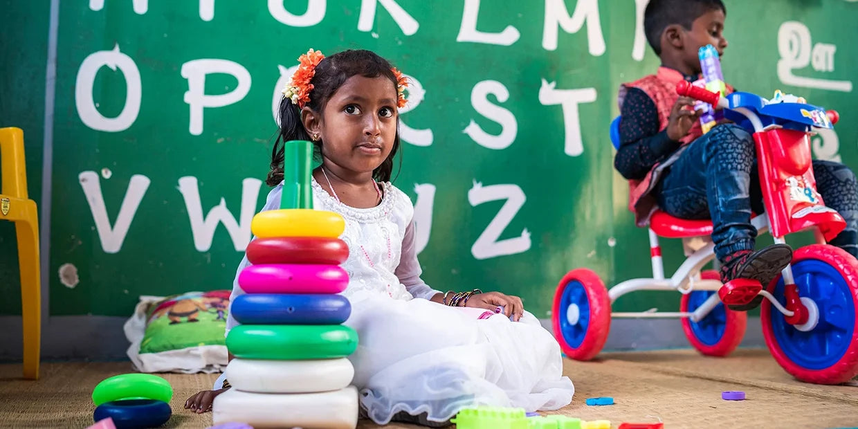 Ein Kind sitzt im Kindergarten auf dem Boden und spielt mit buntem Spielzeug. Im Hintergrund ist eine grüne Wand mit aufgemalten Buchstaben.