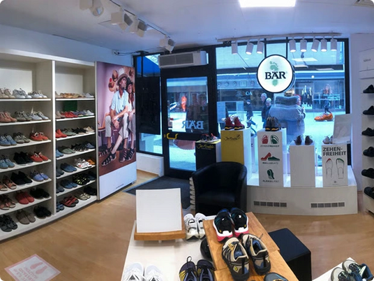 Innenansicht eines modernen Schuhladens mit diversen Sneakern und Lederschuhen in Regalen, samt ‚Zehenfreiheit‘-Plakat an der Wand