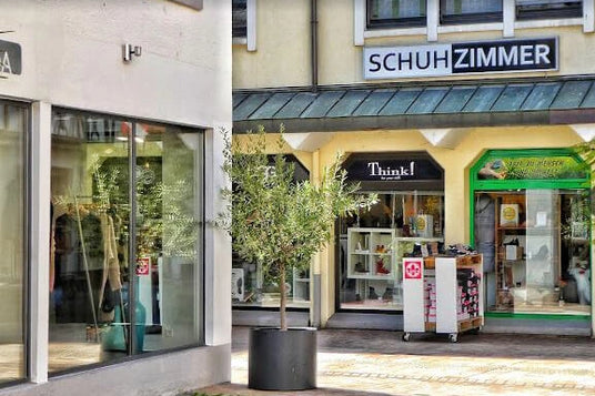 Foto eines kleinen Einkaufsbereichs mit Schaufenstern und dem Eingang eines Schuhgeschäfts namens Schuhzimmer. Es gibt einen Olivenbaum im Topf vor dem Geschäft.